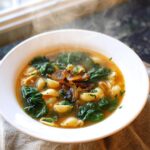 A steaming white bowl of Garlic Ginger Immune Boosting Soup containing shell pasta, dark green leafy vegetables, and caramelized onions.
