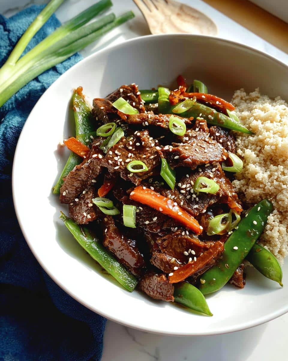 A close-up of a bowl containing Gluten-Free Beef Stir Fry with snap peas, carrots, and sesame seeds, served next to quinoa.