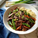 A white bowl filled with Gluten-Free Beef Stir Fry, featuring beef, snow peas, carrots, and sesame seeds, served alongside quinoa.