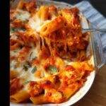 A fork lifts a serving of hot Gluten-Free Chicken Pasta Bake, showing long, melted cheese pulls over tomato sauce and chicken pieces.