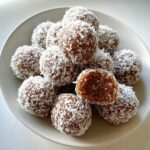 A pile of Gluten-Free Coconut Bliss Balls coated in shredded coconut, with one ball cut open showing the dense interior.