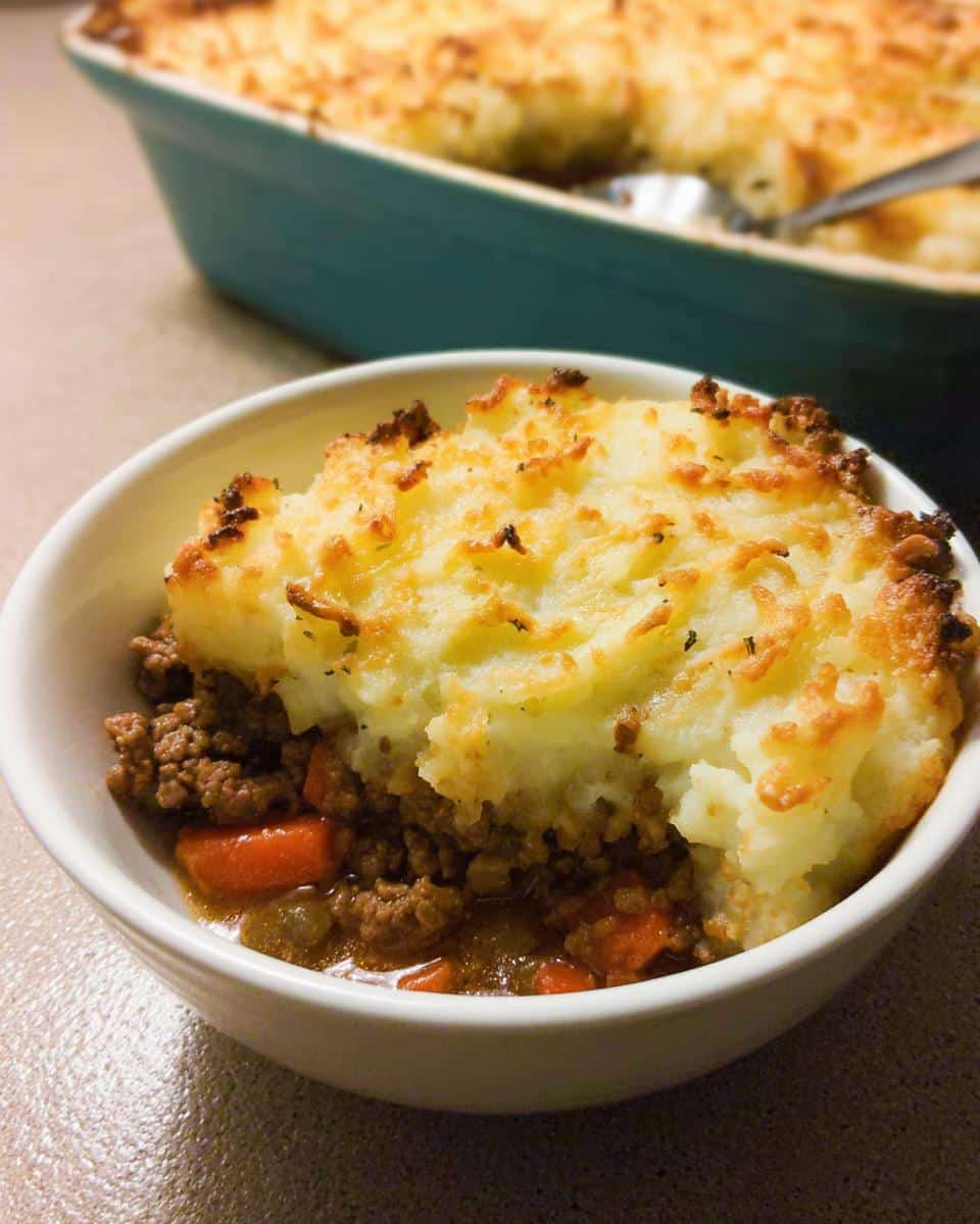 A single serving of Gluten-Free Shepherd’s Pie in a white bowl, showing ground meat and carrots topped with browned mashed potatoes.
