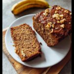 A slice and a small loaf end of moist Gluten-Free Vegan Banana Bread topped with walnuts on a white plate.