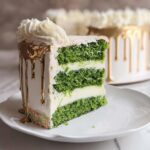A tall slice of Green Velvet Cake with vibrant green layers, white frosting, and gold drip icing.