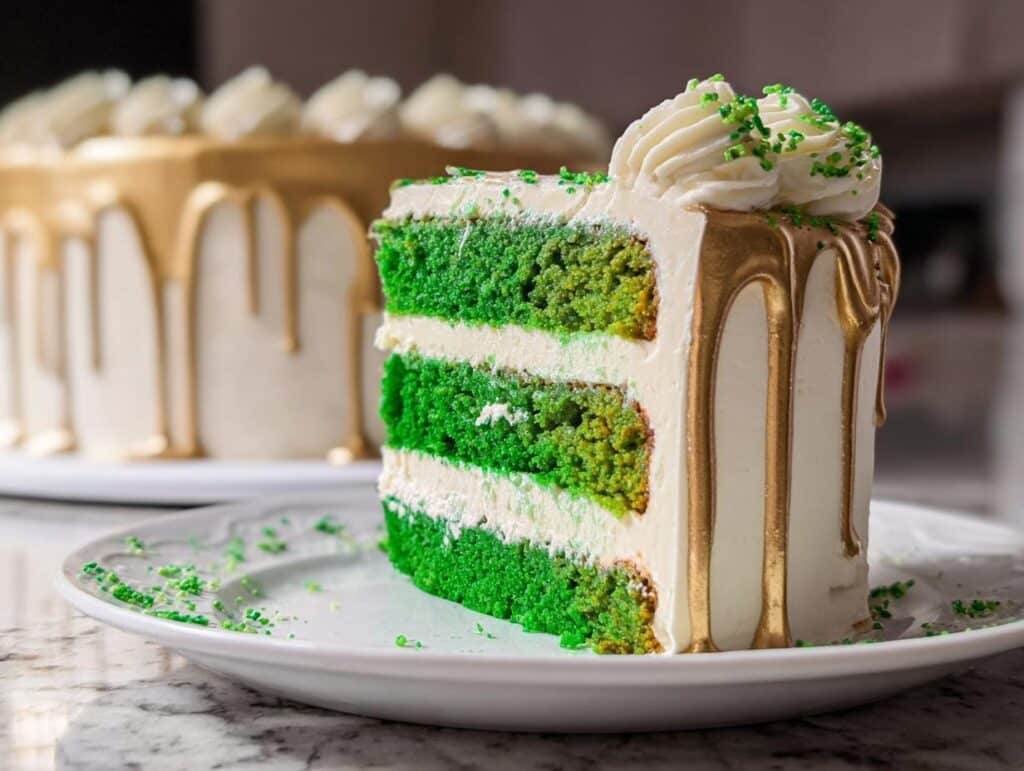 A tall slice of Green Velvet Cake featuring three vibrant green layers, white frosting, and a striking gold drip effect.