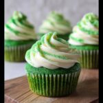 A close-up of a vibrant Green Velvet Cupcake topped with white frosting and green sprinkles, sitting on a wooden board.