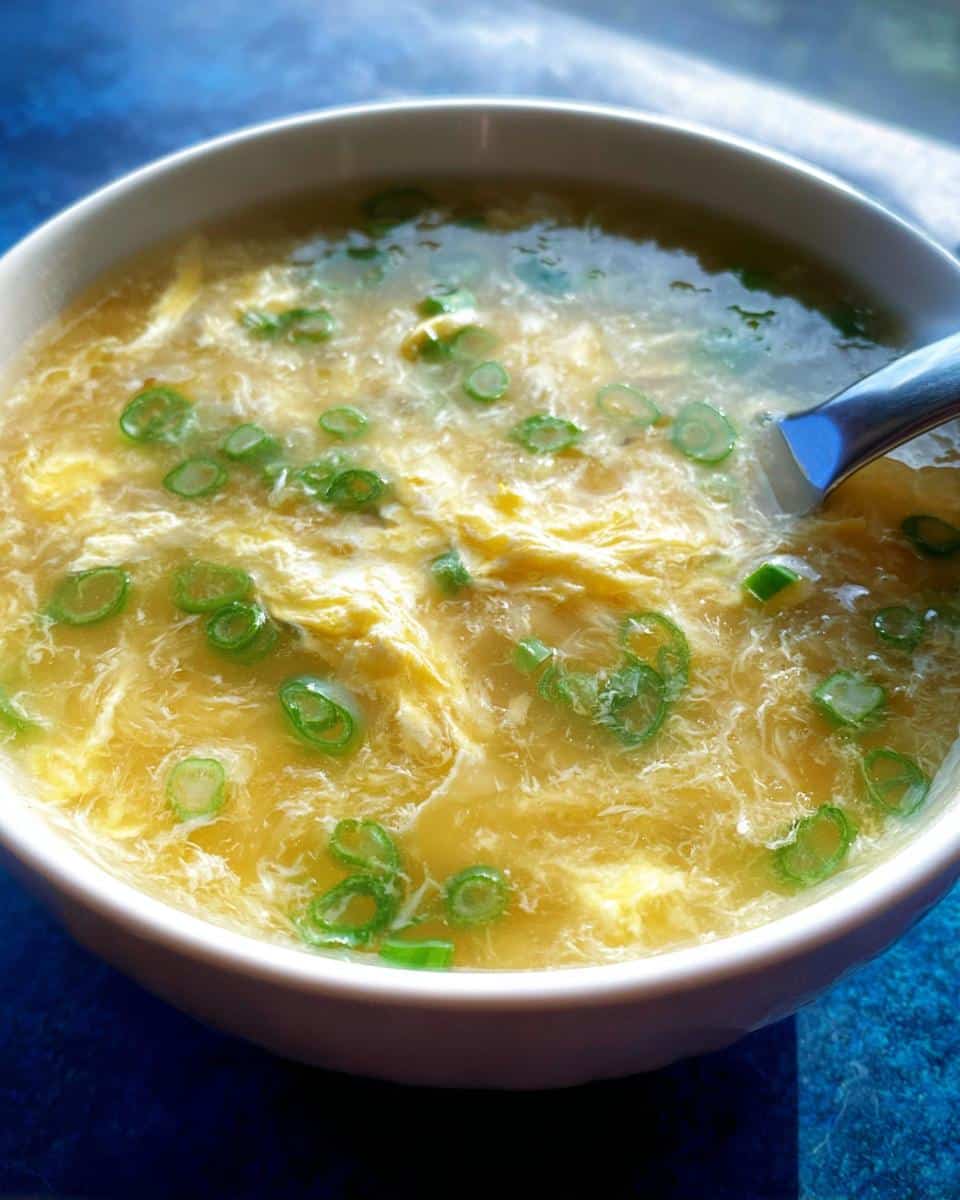 Close-up of a bowl of healthy Egg Drop Soup featuring wispy egg ribbons and bright green scallion slices.