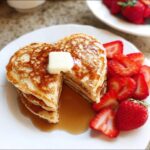 Stack of golden Heart Pancakes Valentine Breakfast topped with butter and syrup, served with fresh sliced strawberries.