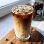 A tall glass of Iced Vanilla Latte Starbucks Style showing layers of milk, espresso, and ice cubes, sitting on a wooden board.