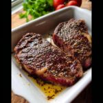 Two thick-cut steaks resting in a white dish covered in a rich, herby Keto Steak Marinade.