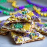A stack of King Cake Cracker Candy pieces topped with white frosting and purple, green, and gold sanding sugar.