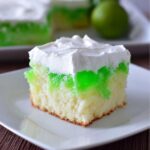 A square slice of moist white cake soaked with bright green gelatin, topped with whipped cream, served on a white plate, featuring Lime Poke Cake.