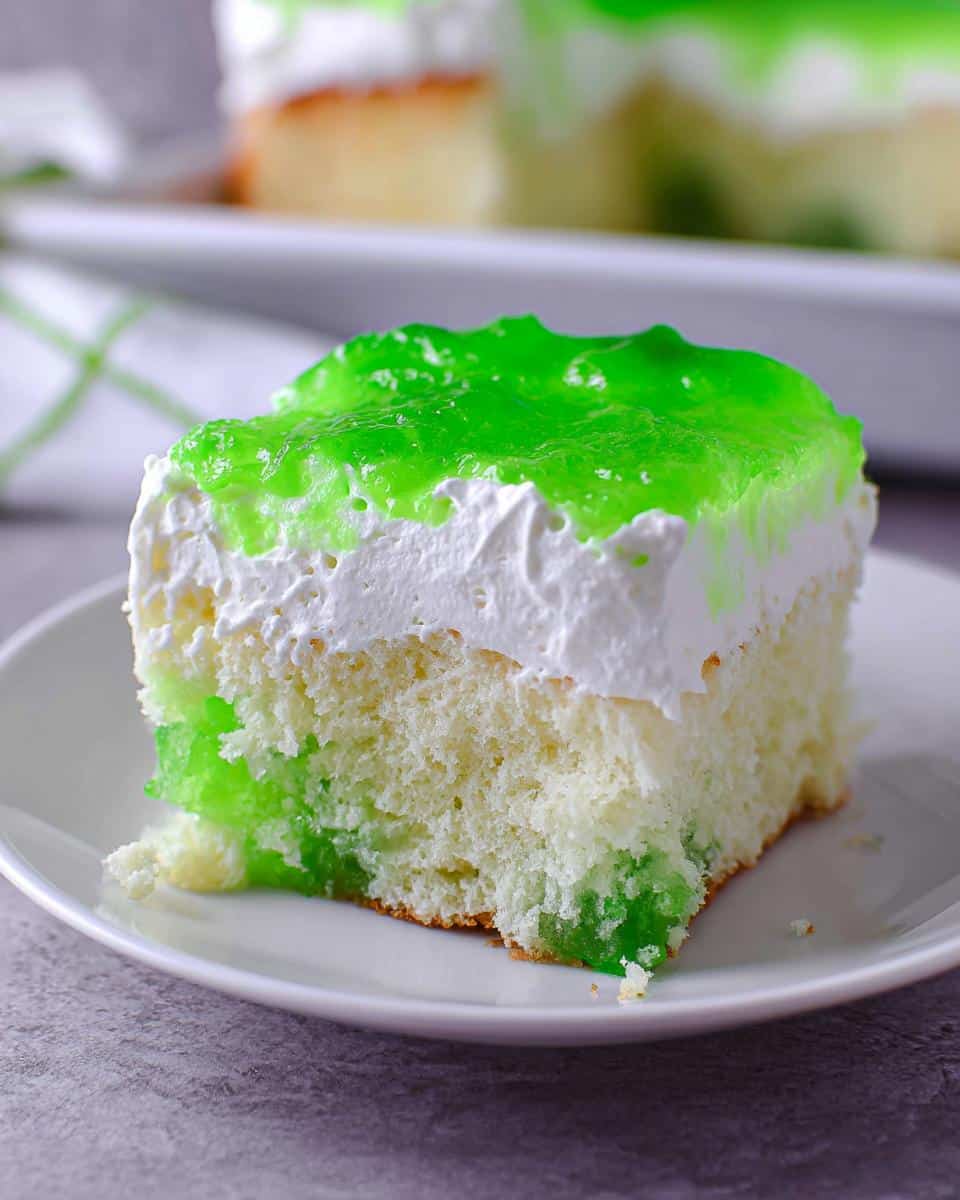 A moist slice of Lime Poke Cake showing the white cake base soaked with bright green lime gelatin and topped with whipped cream.