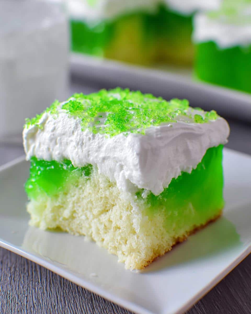 A square slice of moist Lime Poke Cake featuring bright green gelatin, white whipped topping, and green sprinkles.