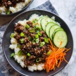 A close-up of a Low Carb Korean Beef Bowl featuring ground beef over white cauliflower rice, topped with green onions and sesame seeds.