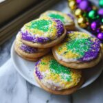 A stack of round Mardi Gras Cake Mix Cookies topped with white icing and purple, green, and yellow sprinkles.