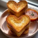 Two golden brown Mini Grilled Cheese Hearts stacked on a pink plate next to a small bowl of tomato soup.