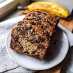 Close-up of several thick slices of Moist Chocolate Chip Banana Bread stacked on a light plate, with a whole banana blurred in the background.