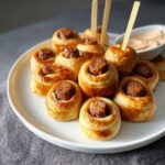 A plate of golden brown New Orleans Style Sausage Bites wrapped in puff pastry, served with toothpicks.