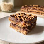 Two stacked No-Bake Chocolate Oat Bars topped with a rich chocolate layer and drizzled with caramel sauce.