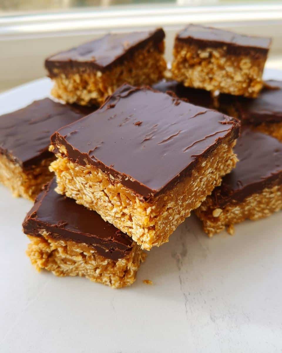 Close-up of several square No-Bake Peanut Butter Oat Bars stacked on a white surface, topped with smooth chocolate.