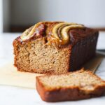 A close-up of a sliced Oil-Free Vegan Banana Bread loaf topped with caramelized banana slices.