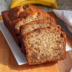 Close-up of thick slices of moist One-Bowl Easy Banana Bread on a white platter, with a whole banana blurred in the background.