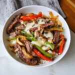 Close-up of a Philly Cheesesteak Protein Bowl featuring steak, peppers, onions, mushrooms, and melted white cheese over rice.