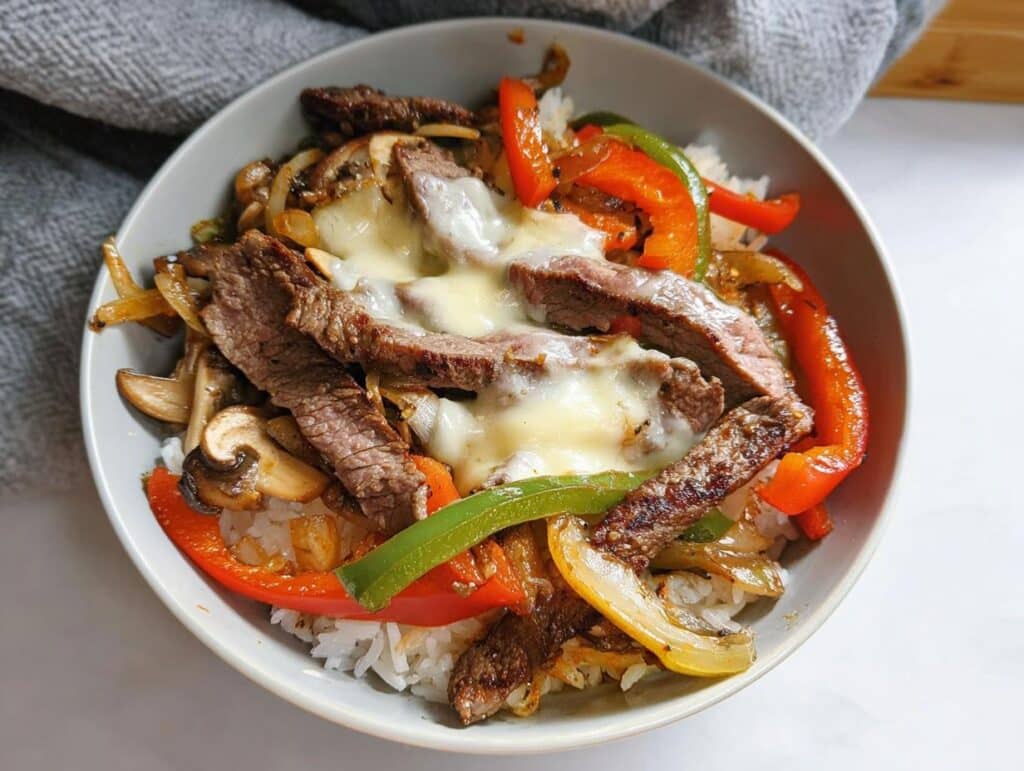 A close-up of a Philly Cheesesteak Protein Bowl featuring sliced steak, peppers, onions, mushrooms, and melted cheese over white rice.