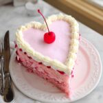 A slice of moist Pink Heart Cake with pink frosting, white piped edges, and a maraschino cherry on top.