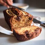 A rich, moist slice cut from a Plant-Based Banana Bread Loaf topped with banana and chocolate chips.
