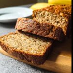 Two thick slices of moist Protein Banana Bread resting against the main loaf on a wooden cutting board.