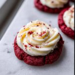 A close-up of a vibrant Red Velvet Cupcake Cookie topped with a swirl of cream cheese frosting and red sprinkles.