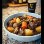 A close-up of a bowl of rich Rustic Beef Stew with chunks of beef, carrots, and potatoes, with a pint of Guinness in the background.