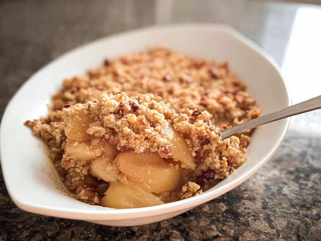 A spoonful being taken out of a warm serving of Gluten-Free Apple Crumble, showing soft apples and crunchy pecan topping.