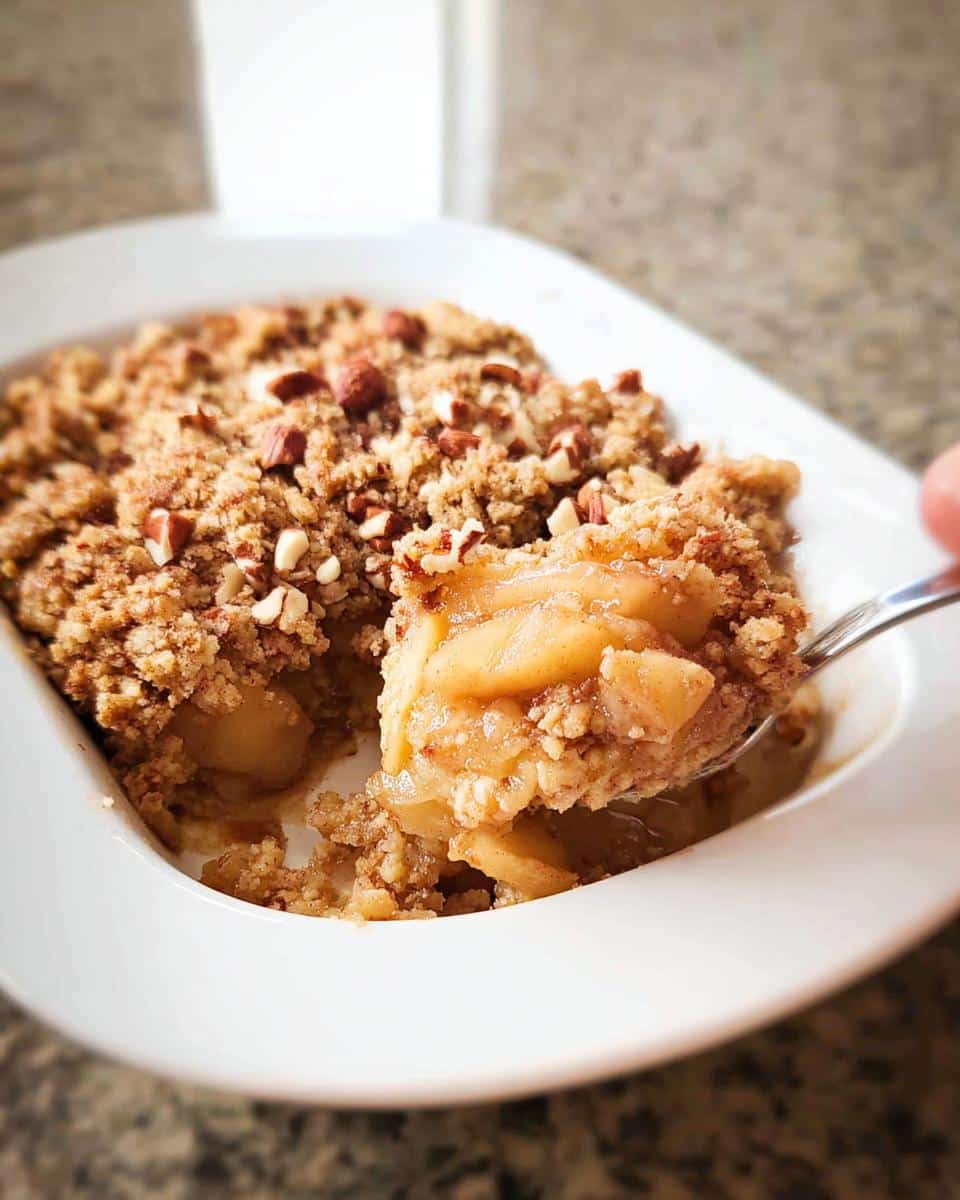 A spoonful of warm Gluten-Free Apple Crumble being lifted from a white dish, showing soft apples and crunchy topping.