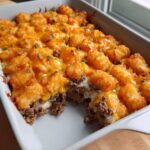 A close-up of a Simple and Delicious Tater Tot Casserole with a scoop removed, showing layers of ground beef, creamy sauce, and crispy tater tots topped with melted cheddar.