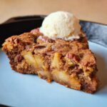 A close-up of a warm slice of Skillet Apple Brownie topped with a scoop of melting vanilla ice cream.