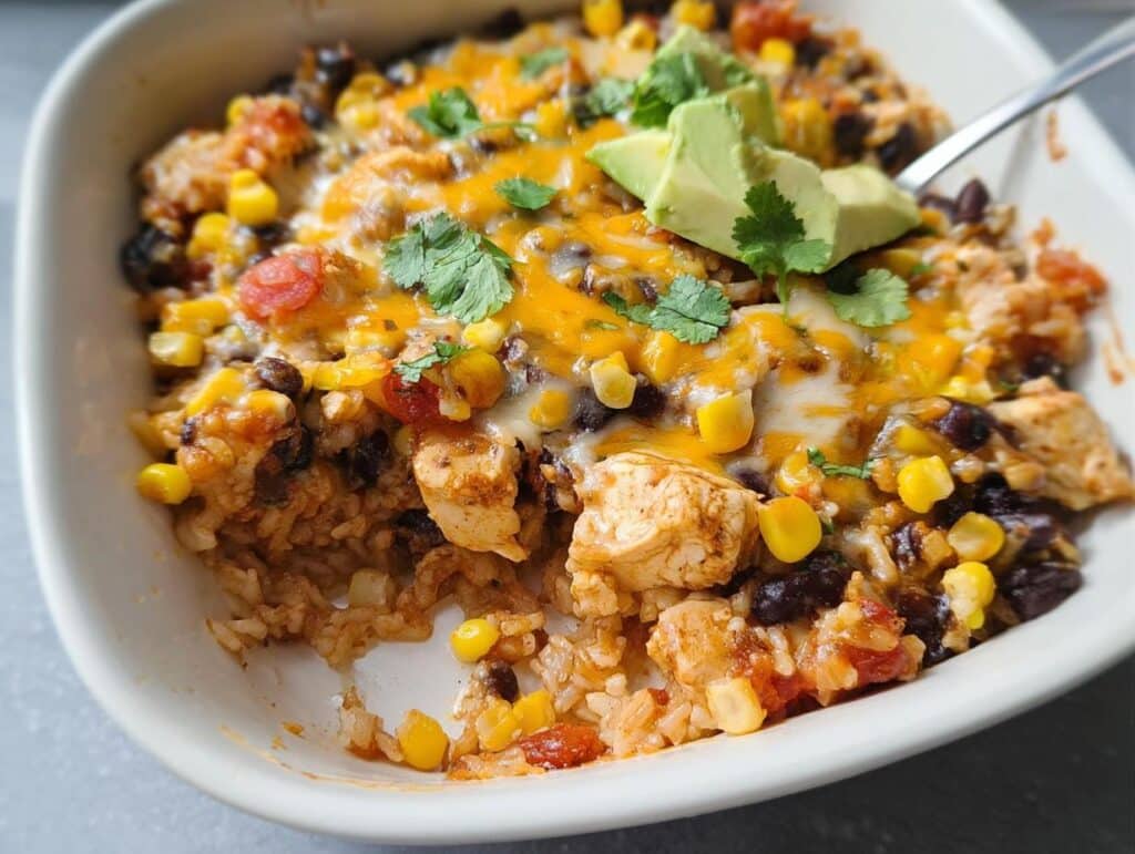 A close-up of a scoop taken from a Southwest Chicken Casserole topped with melted cheese, corn, beans, and avocado.