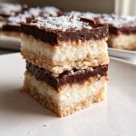 Two stacked squares of No-Bake Coconut Dessert Bars featuring a crust, thick coconut layer, and chocolate ganache, topped with shredded coconut.