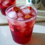 Close-up of a vibrant red Strawberry Acai Refresher Copycat drink filled with ice cubes and topped with freeze-dried strawberries.