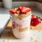 A layered Strawberry Cheesecake Overnight Oats dessert served in a glass jar, topped with fresh strawberries and granola.
