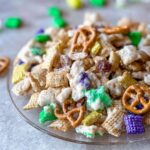 A close-up of a sweet Mardi Gras Snack Mix featuring cereal, pretzels, and colorful green, purple, and gold coated candies.