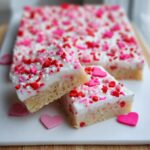 Two squares of Valentine Sugar Cookie Bars topped with white frosting and red/pink heart sprinkles, stacked slightly.