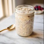 A jar filled with creamy Vanilla Cream Overnight Oats, topped with rolled oats, sitting on a marble surface.