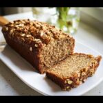 A loaf of moist Vegan One-Bowl Banana Bread, partially sliced, topped with oats.