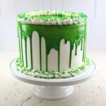 A tall, white frosted cake featuring a vibrant green drip glaze and green sprinkles, showcasing a perfect Green Drip Cake.