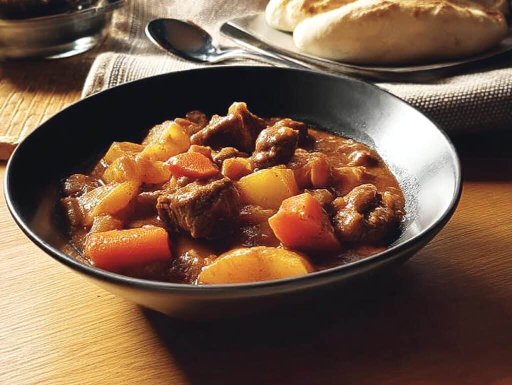 A close-up of a dark bowl filled with rich Winter Beef Stew chunks, potatoes, and carrots, served on a wooden table.