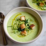 A bowl of vibrant green Zucchini Basil Soup topped with zucchini noodles, basil leaves, and golden croutons.